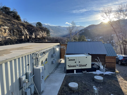 A Newterra Clear3 decentralized membrane bioreactor system in the mountains of Telluride, Colorado with the system consisting of a white shipping container with double doors, electrical panels, and meters with electrical power hooked up via a Kohler branded power unit with a small municipal building and parking area