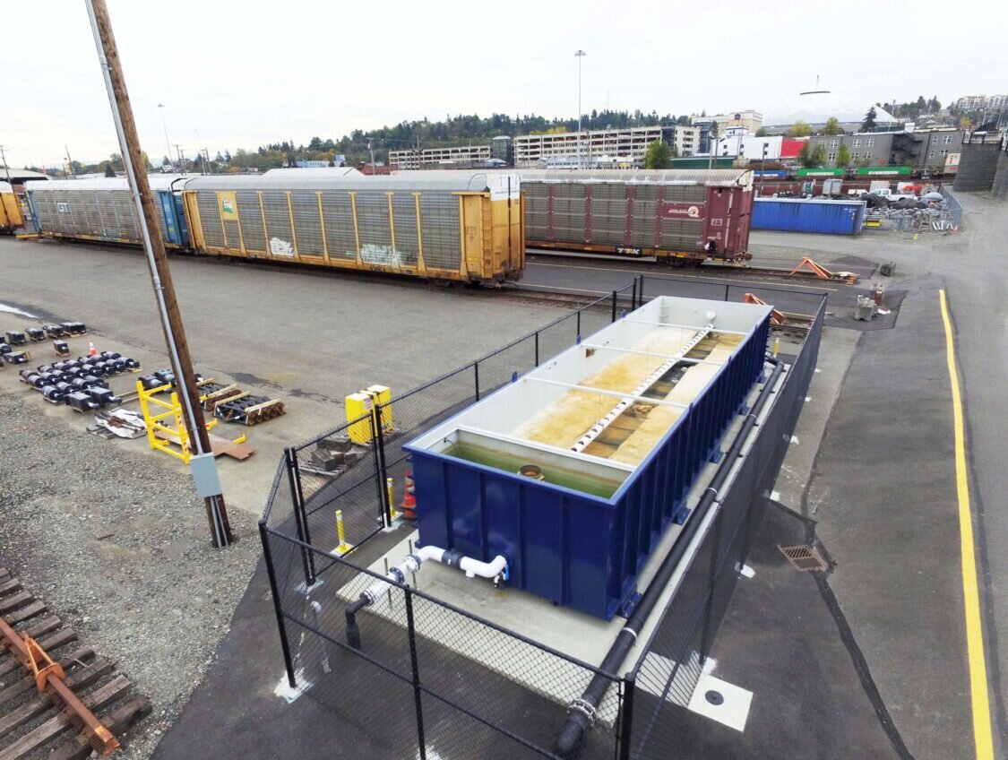 Low aerial view of a railyard with a blue Newterra modular water treatment processing unit divided into two bays connected to an underground pipe with a chain link fence around the unit, train cars sit nearby on the end of the rail line, condos and apartment buildings of a medium size city are in the background