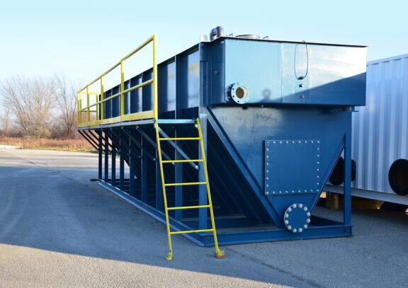 A Newterra Clara longbox filtration unit in an industrial setting consisting of a yellow ladder and guardrail attached to the unit with a V shaped base running the length of the box with a rectangular box over it, riveted access panels can be seen at the base of the unit