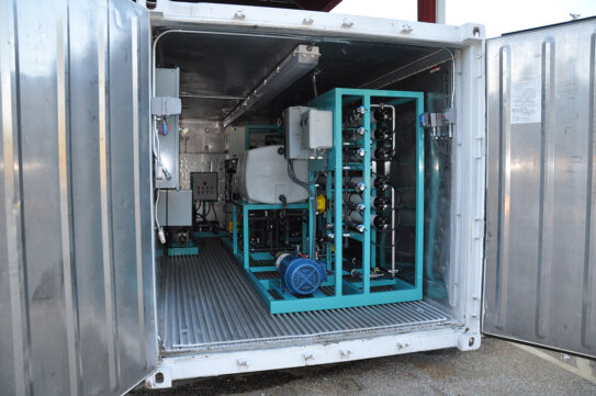 A modular mobile Seamega sea water desalination system consisting of a shipping containing housing a cyan metal frame supporting a small motor, water tank, electrical panels, and various horizontally positioned filtration pipe units, overhead lighting and additional electrical panels accompany the unit