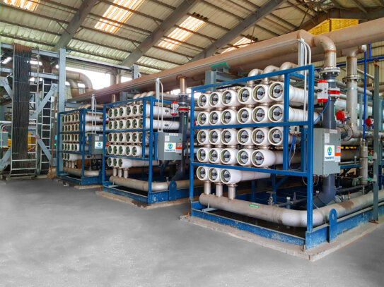 Three reverse osmosis Delta units installed on a concrete slab inside an industrial warehouse with each unit consisting of horizontally oriented plastic filtration pipe units stacked within a blue metal frame along with an electrical panel and pipes of various sizes with valve and gauge components