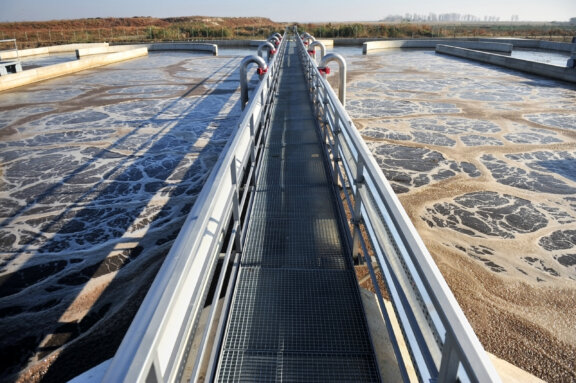 Close up of a Newterra Tri Oval oxidation ditch wastewater treatment system including a grated metal walkway running the length of the system with water being treated on either side underneath the walkway, concrete divisions in the system are present on both sides of the walkway with trees in the background within a rural outdoor setting