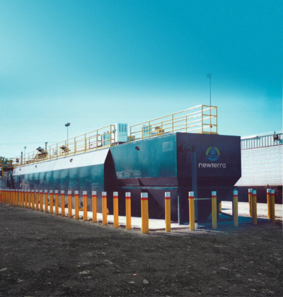 Aerial view of a Newterra Unisystem package wastewater system with a maintenance walkway across the top of the main aeration tanks and two clarification chambers, various upright metal containers and control panels are placed to the left of the treatment unit in an outdoor environment, a streetlight is on the right