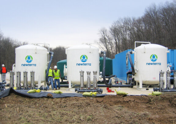 Three technicians inspect three Newterra branded tanks used for treating wastewater in an outdoor setting surrounded by trees