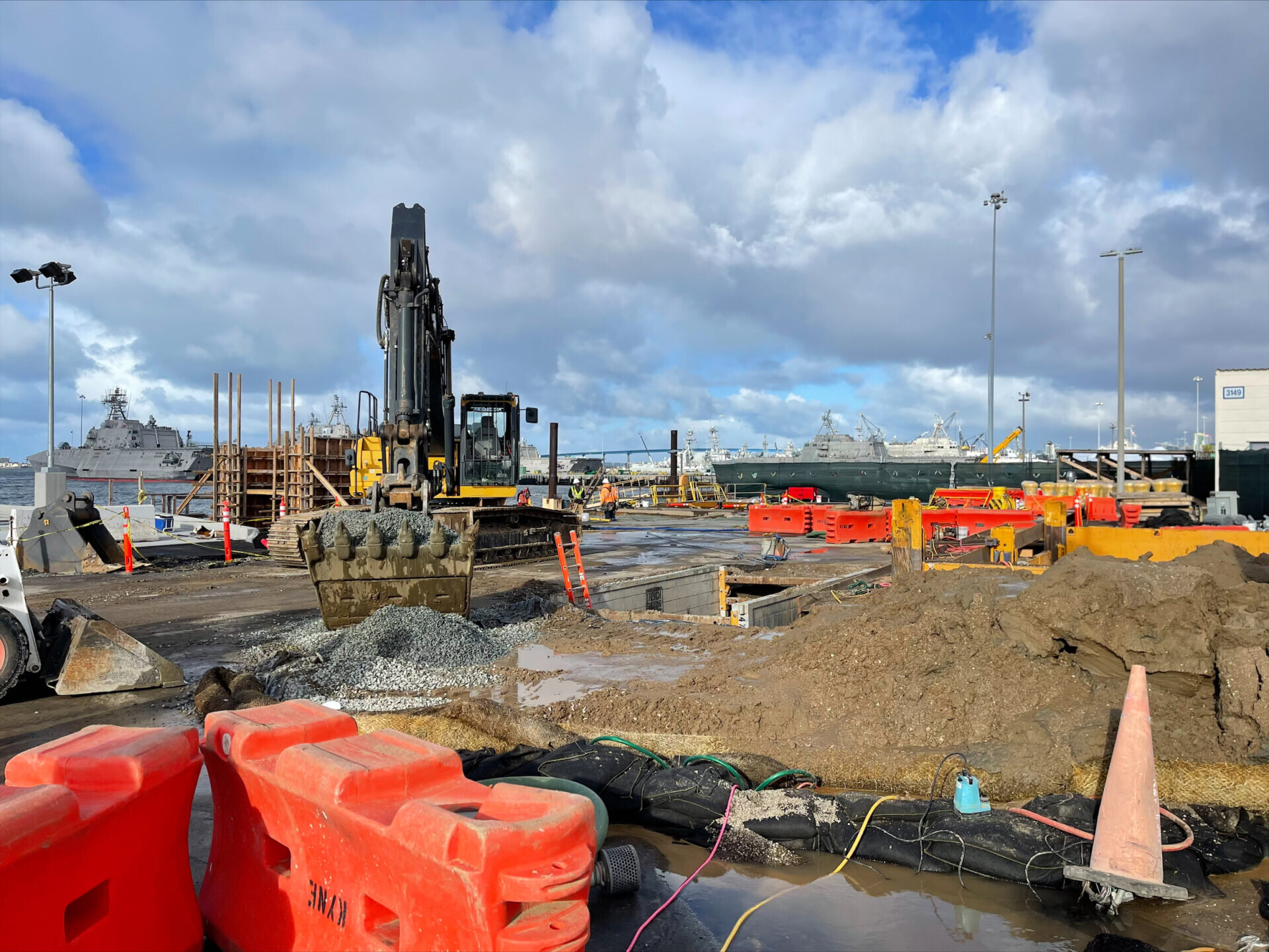 View of a large port with a military ships docked near a yard with a scoop excavator on tracks removing gravel material next to a pile of earth and sand