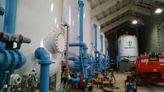 View from inside a water treatment containing six water treatment tanks connected with source or output pipes extending away from the tanks in a brick building