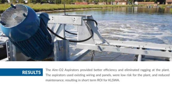 Image with caption of Aire-O2 aspirator in use at an open air pond with grass and single story industrial or commercial brick buildings in the background, foreground contains close up of equipment aerating the pond water