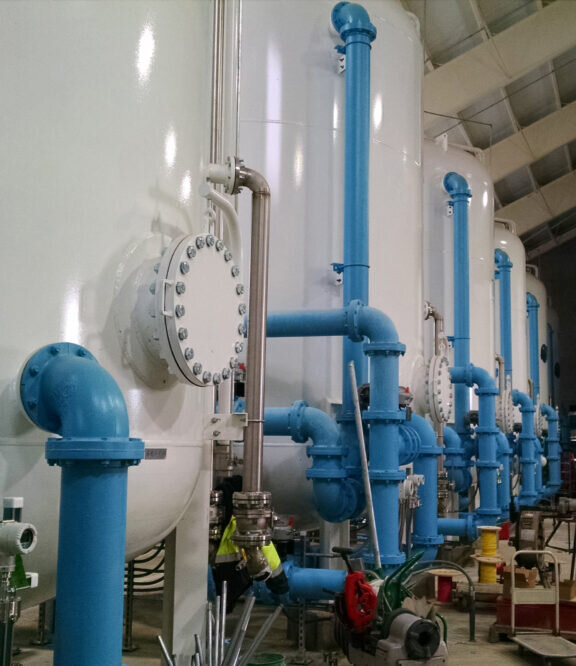 View from inside a water treatment containing six water treatment tanks connected with source or output pipes extending away from the tanks in a brick building