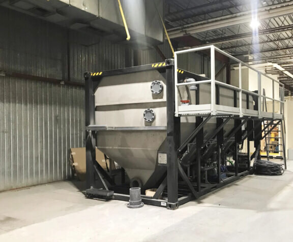 A Newterra longbox filter sits in an industrial building consisting of a metal box with a v shaped base with a black frame and a maintenance access walkway and set of stairs